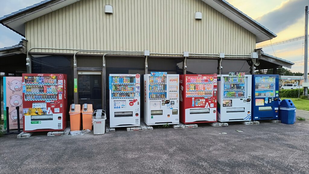 コカ・コーラ、DyDo、伊藤園など複数メーカーの自動販売機が6台並ぶ列。手前には缶・瓶用とペットボトル用のゴミ箱も設置されている。
